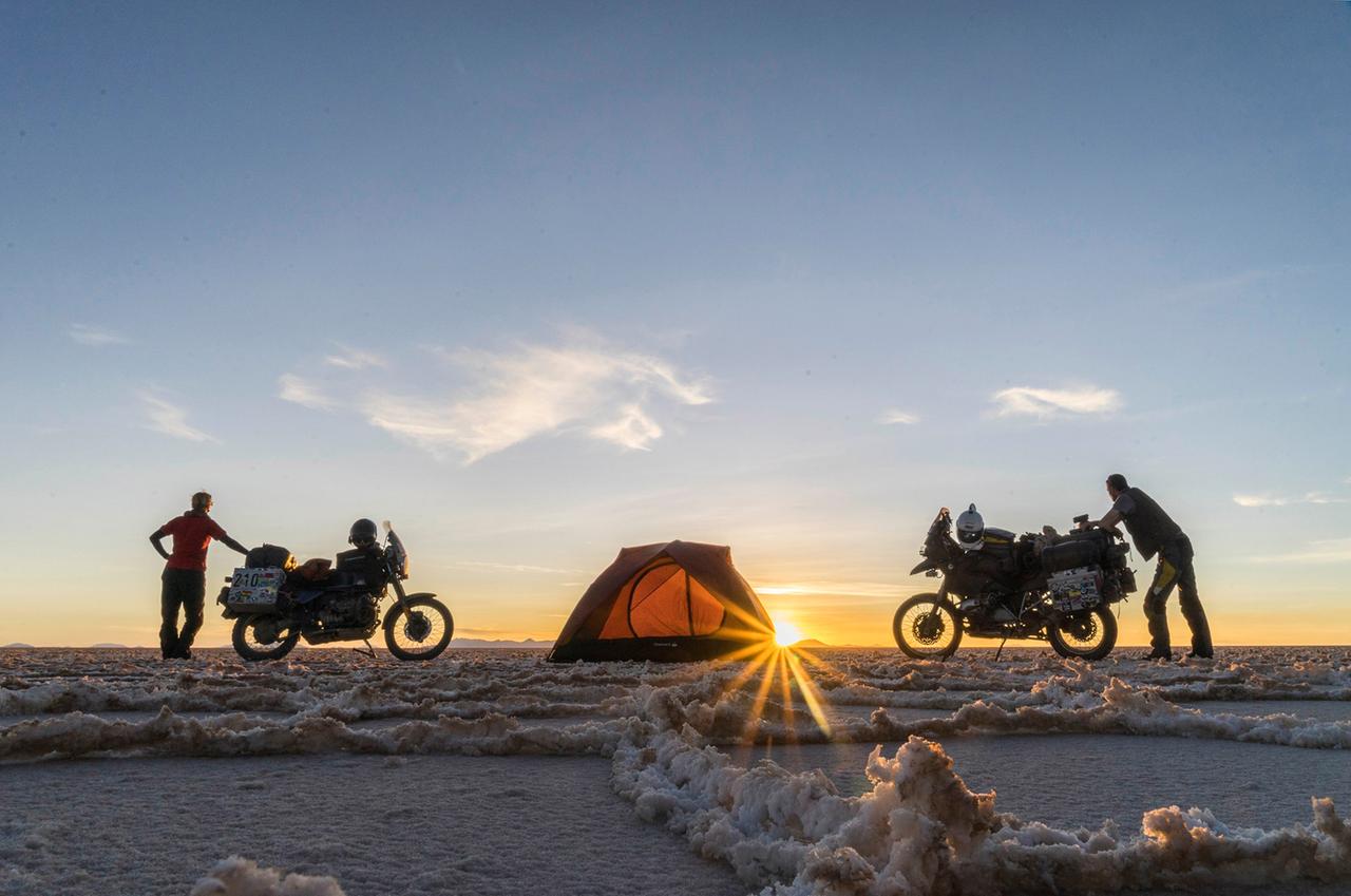 Zwei Menschen stehen bei Sonnenaufgang neben ihren Motorrädern, in der Mitte steht ihr Zelt.